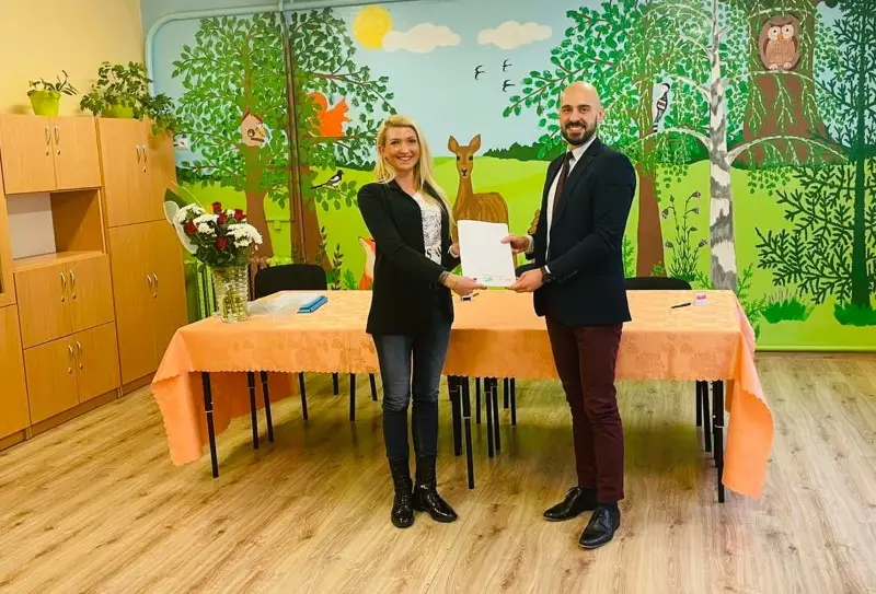 Zdjęcie o orientacji poziomej przedstawia dwie osoby stojące w sali przedszkolnej i trzymające wspólnie biały dokument. Kobieta po lewej stronie ubrana jest w czarny żakiet, jeansy i czarne botki, natomiast mężczyzna po prawej ma na sobie elegancki ciemny garnitur z bordowymi spodniami. Postacie znajdują się przed stołem nakrytym pomarańczowym obrusem, na którym stoi wazon z białymi i czerwonymi kwiatami. Tło stanowi kolorowy mural z motywem leśnym, na którym widoczne są drzewa, sarna, wiewiórka i sowa. Po lewej stronie kadru znajdują się jasne, drewniane szafki w zabudowie, a na podłodze ułożone są panele imitujące drewno, co nadaje wnętrzu przytulny, edukacyjny charakter.