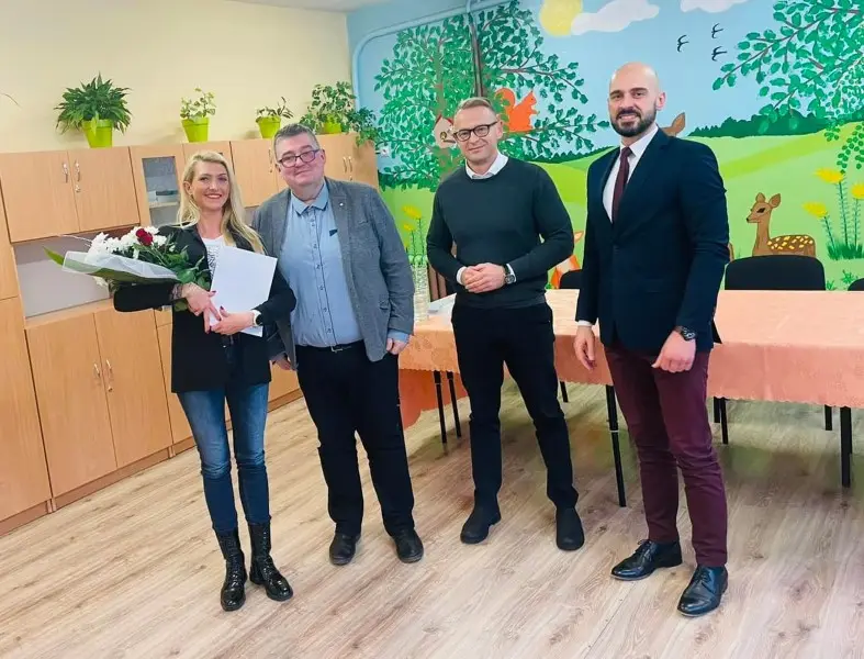 Zdjęcie w orientacji pionowej przedstawia grupę czterech osób stojących w jasnej sali, prawdopodobnie w przedszkolu lub szkole podstawowej. Po lewej stronie stoi kobieta trzymająca bukiet kwiatów oraz dokument, a obok niej pozuje trzech mężczyzn ubranych w stylizacjach od półformalnych po eleganckie. W tle widać drewniane szafki, stół przykryty brzoskwiniowym obrusem oraz kolorowy mural na ścianie przedstawiający leśne zwierzęta i drzewa. Podłoga w pomieszczeniu wyłożona jest jasnymi panelami, co nadaje wnętrzu ciepły i przytulny charakter. Cała kompozycja jest ustawiona frontalnie, co podkreśla oficjalny charakter spotkania lub uroczystości.