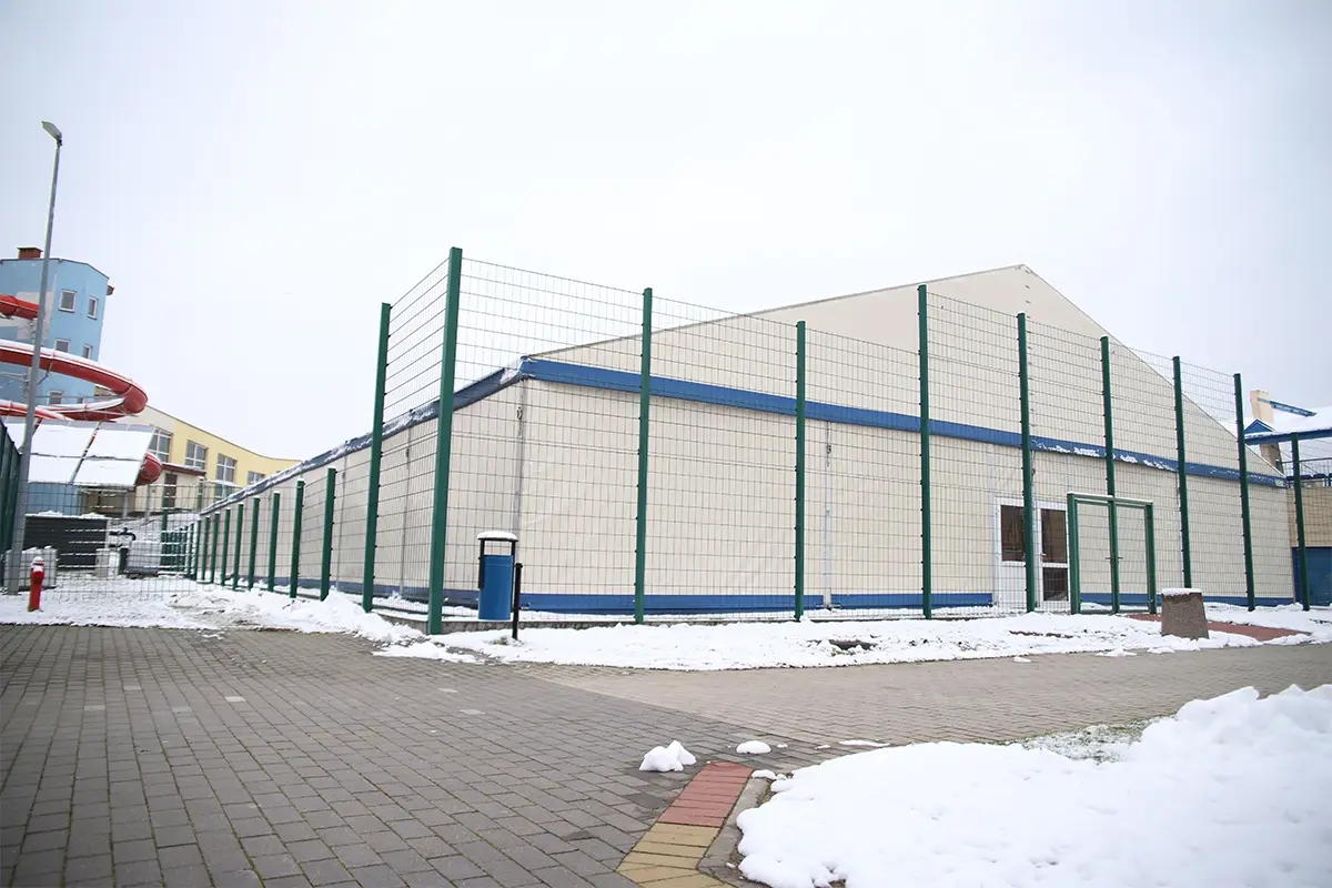 Zdjęcie przedstawia dużą halę namiotową o jasnych ścianach z niebieskimi akcentami, otoczoną wysokim zielonym ogrodzeniem z siatki. Obiekt o proporcjach boków zbliżonych do kwadratu położony jest na wybrukowanym placu, na którym widać zalegający śnieg. W lewej części kadru widoczny jest fragment niebieskiego budynku z czerwoną rurą zjeżdżalni oraz metalowy słup oświetleniowy. Na pierwszym planie po lewej stronie znajduje się niebieski kosz na śmieci, a podłoże wyłożone jest betonową kostką brukową. Całość kompozycji ma orientację poziomą i ukazuje infrastrukturę sportową w zimowej scenerii przy pochmurnym niebie.