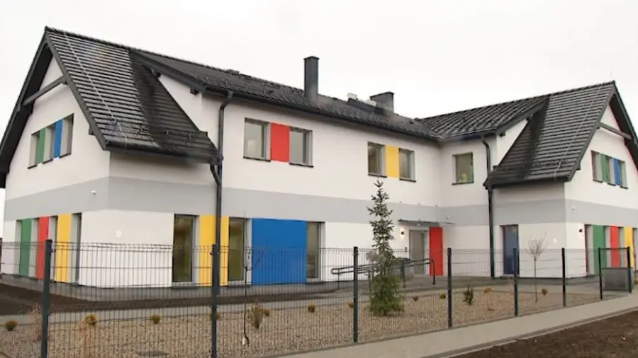 Zdjęcie przedstawia nowoczesny budynek użyteczności publicznej w formacie poziomym, charakteryzujący się jasną elewacją z wyrazistymi, kolorowymi akcentami w odcieniach czerwieni, niebieskiego, żółtego i zielonego. Obiekt posiada dwuspadowy dach pokryty ciemną dachówką oraz szary pas oddzielający kondygnacje, co nadaje mu geometryczny i uporządkowany wygląd. Całość otoczona jest metalowym ogrodzeniem panelowym, za którym znajduje się niewielki teren zielony z młodymi nasadzeniami drzew iglastych. Budynek posiada liczne okna wkomponowane w kolorowe panele ścienne oraz podjazd dla osób niepełnosprawnych prowadzący do głównego wejścia. Dzięki przejrzystej strukturze i kontrastowej kolorystyce, architektura sprawia wrażenie przyjaznej placówki edukacyjnej lub opiekuńczej.